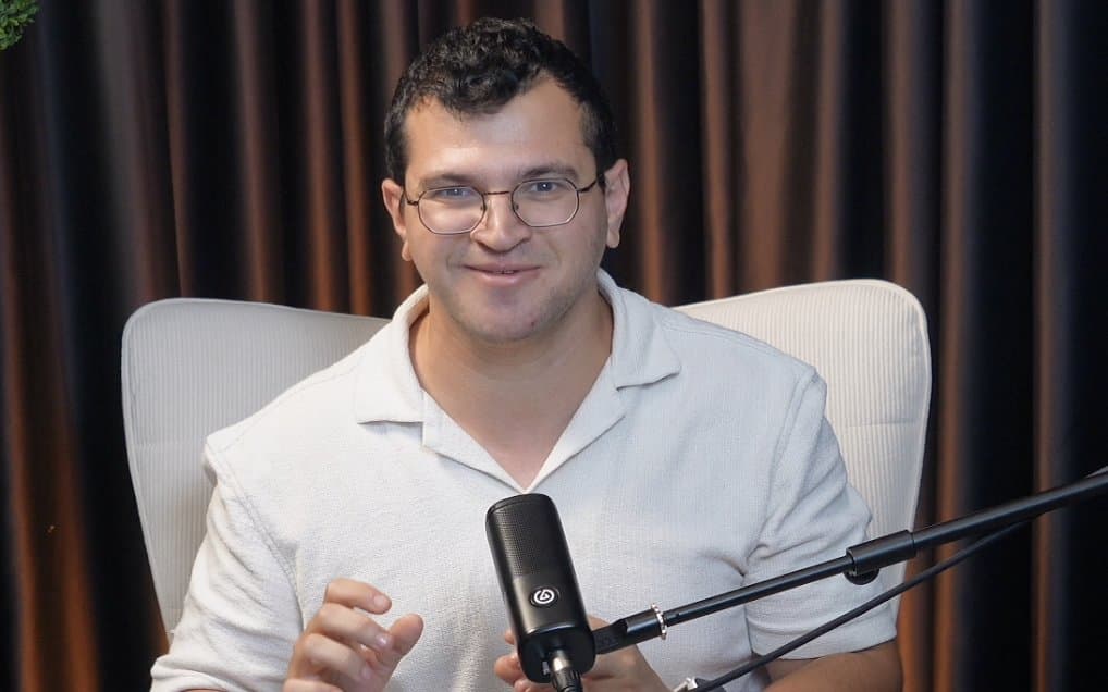 אייל סגל מדבר ישר למצלמה באולפן הבונקר, כורסה לבנה ווילון בתאורה חמה: צילום שיעור לקורס הדיגיטלי שלו